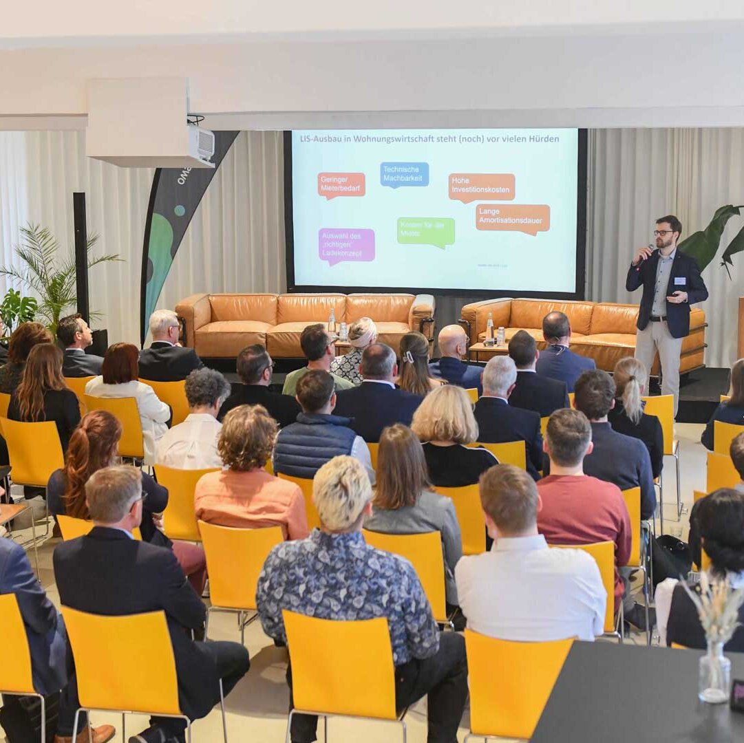 Das Publikum sitzt auf gelben Stühlen und schaut sich eine Präsentation auf einer Leinwand an. 