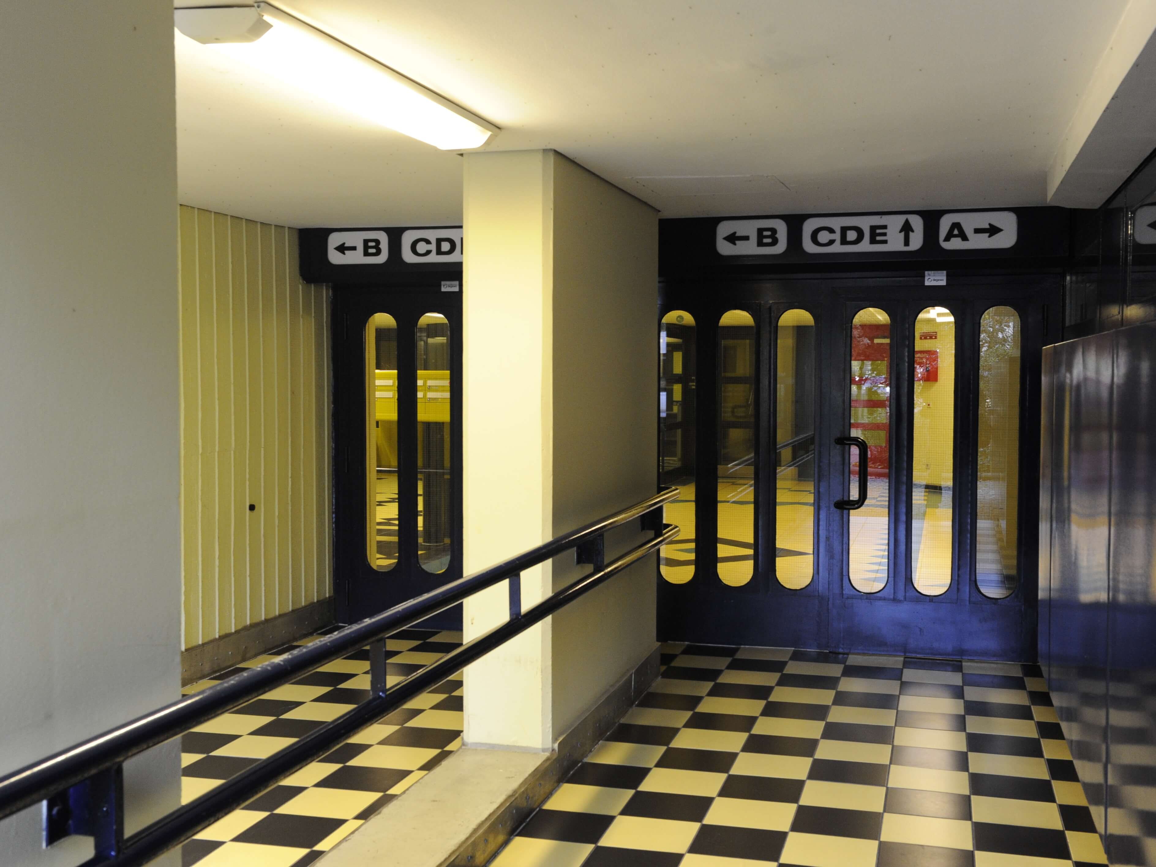 Innenraumzugang der Wohnanlage „Die Schlange“ mit schwarz-weiß kariertem Boden, gelben Wandflächen und blauen Türen mit Wegweisern zu den verschiedenen Gebäudeteilen.