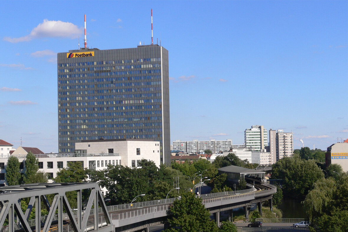 Das ehemalige Postbank-Hochhaus mit dunkler Aluminium-Glas-Fassade, zwei rot-weißen Sendemasten auf dem Dach und dem Postbank-Logo an Außenwand der obersten Etage steht am Halleschen Ufer in Berlin. Vor dem Turm schmiegt sich die U-Bahn-Hochtrasse an den Verlauf des Landwehrkanals.
