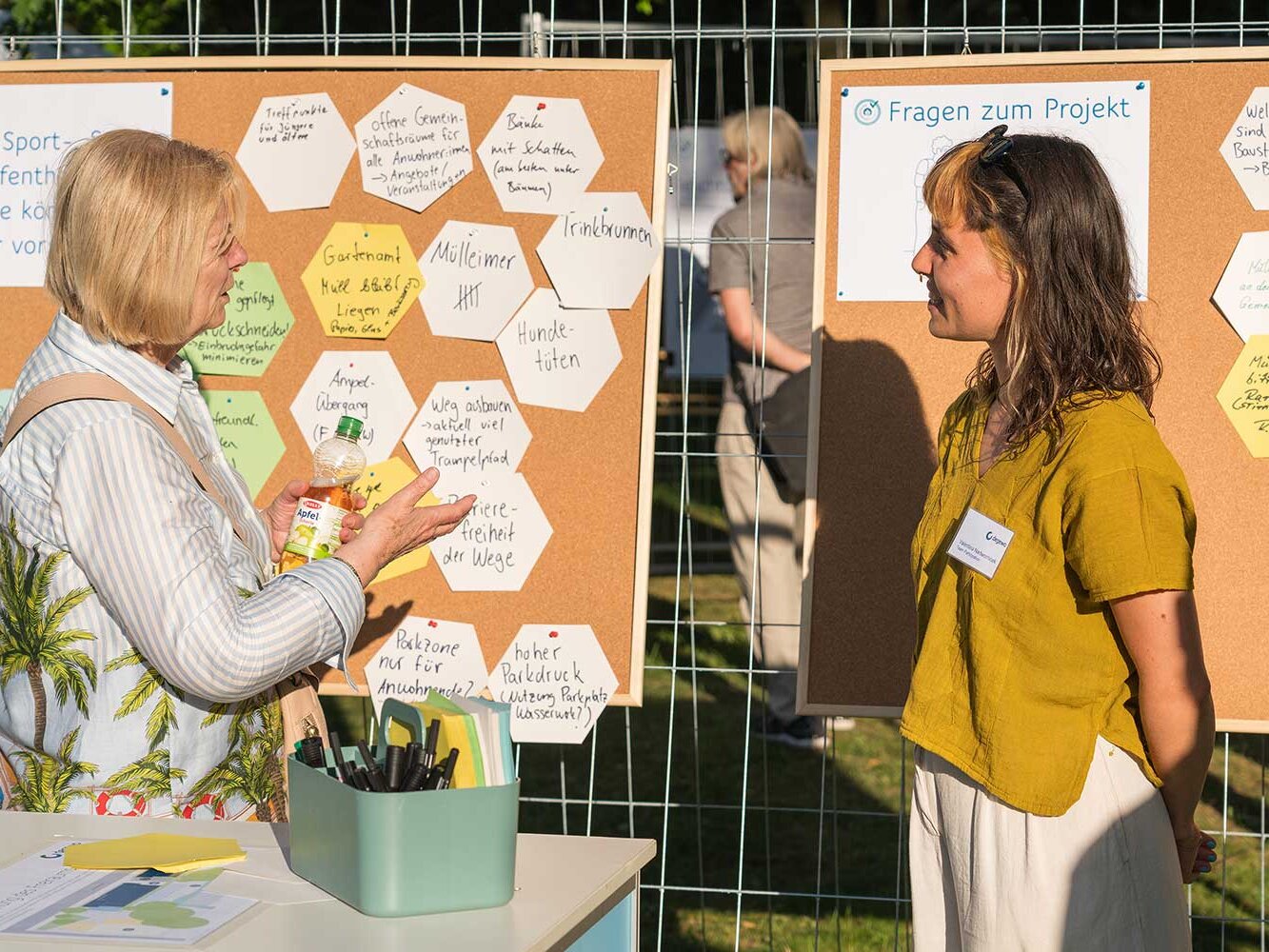 Zwei Frauen stehen im Gespräch vor Pinnwänden mit Fragen und Notizen zum Projekt. Eine Besucherin gibt Feedback, während eine Mitarbeiterin zuhört; im Hintergrund hängen Moderationskarten am Bauzaun.