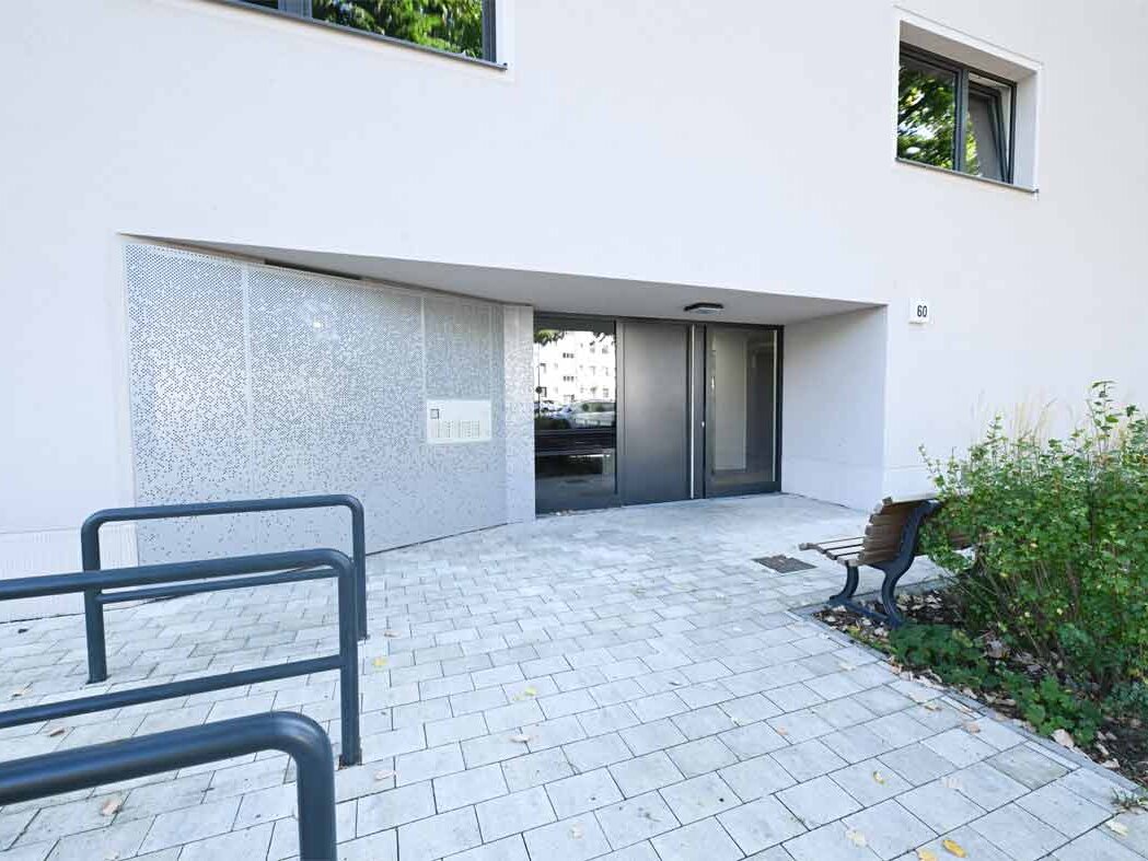 Außenansicht degewo-Neubau Malteserstraße 60: Eingangsbereich mit Fahrradständern links sowie Beet und Sitzbank rechts im Vordergrund.