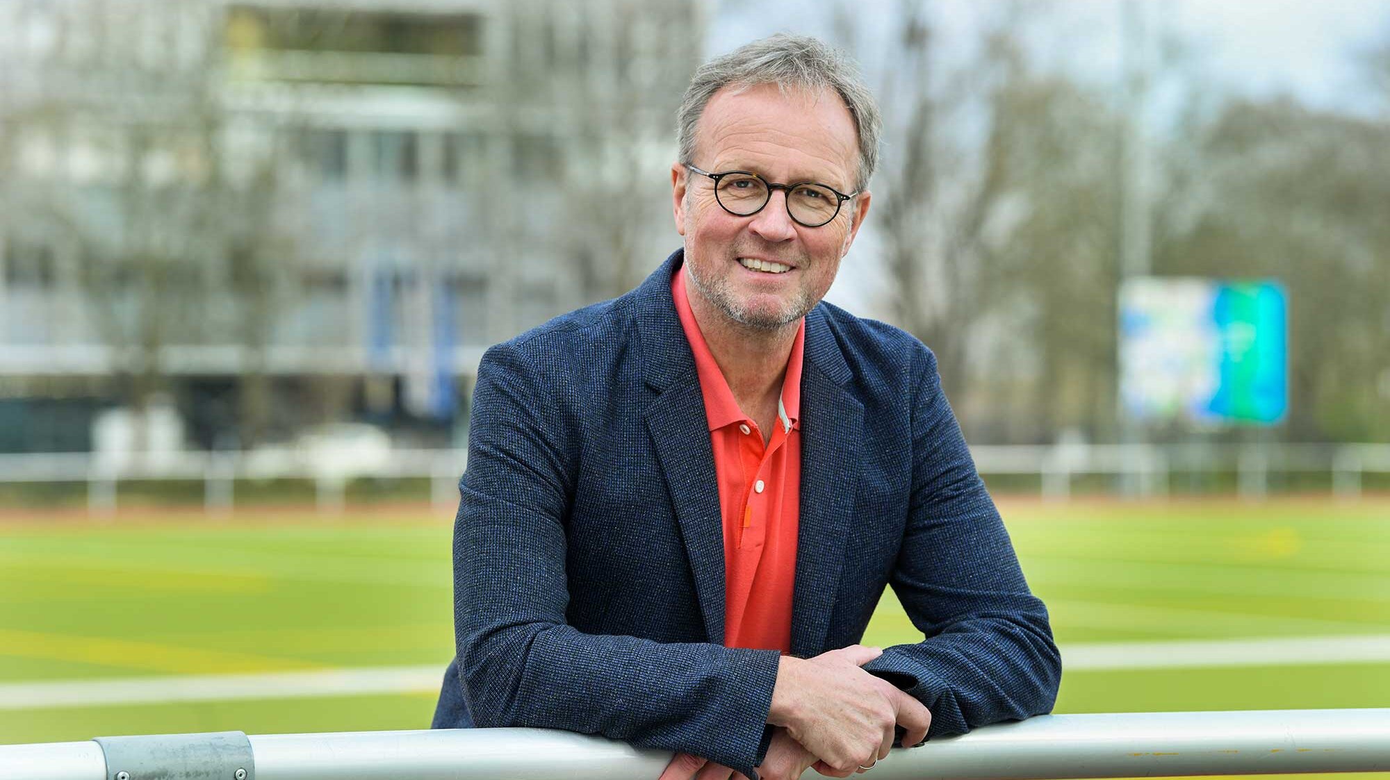 Ein Mann hat seine Arme auf ein Geländer gelegt und lächelt in die Kamera. Im Hintergrund ist verschwommen der Rasen eines Fußballplatzes zu sehen.