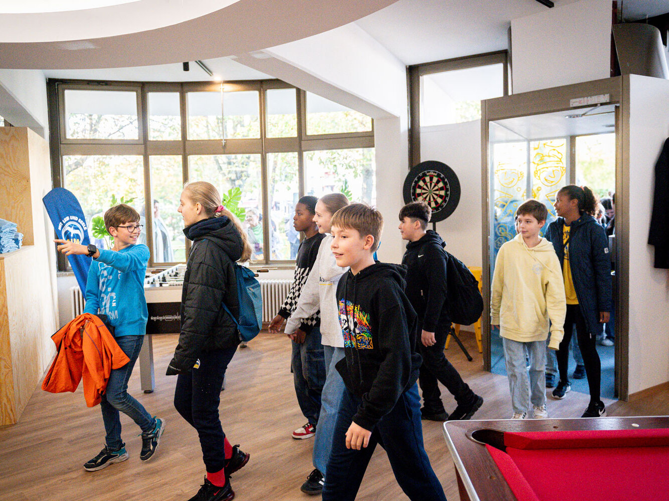 Gruppe von Schulkindern betritt einen hellen Freizeitraum mit Spielangeboten wie Kicker und Darts, während ein Junge stolz ein blaues T-Shirt hochhält.