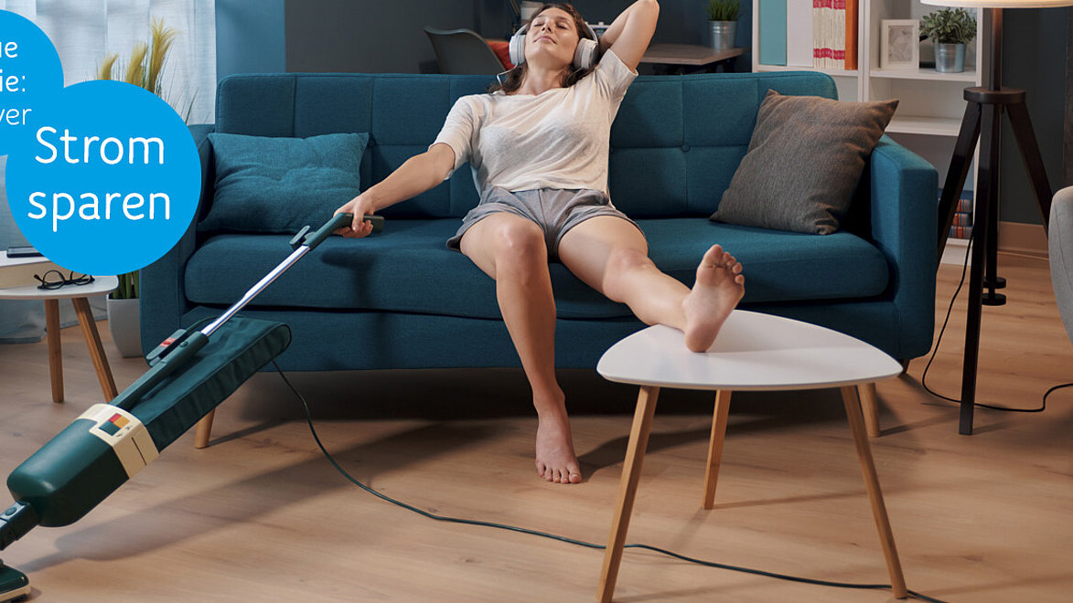 Eine Person sitzt im Wohnzimmer auf dem Sofa. Einen Fuß auf dem Couchtisch, hält sie in der rechten Hand einen Staubsauger und hört dabei über Kopfhörer Musik.