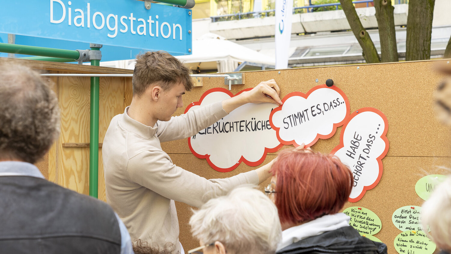 Ein junger Mann befestigt Sprechblasen-Schilder mit Fragen an einer Dialogstation, während mehrere Menschen vor der Pinnwand stehen und zuschauen.