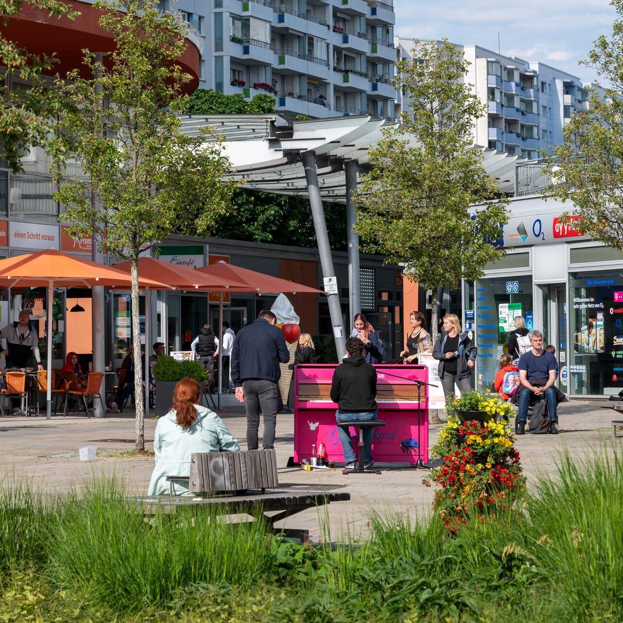 Ansicht eines Teilstückes der Marzahner Promenade. Es sind überdachte Geschäfte zu sehen, auf einem Platz steht ein pinkes Klavier, jemand spielt darauf. 