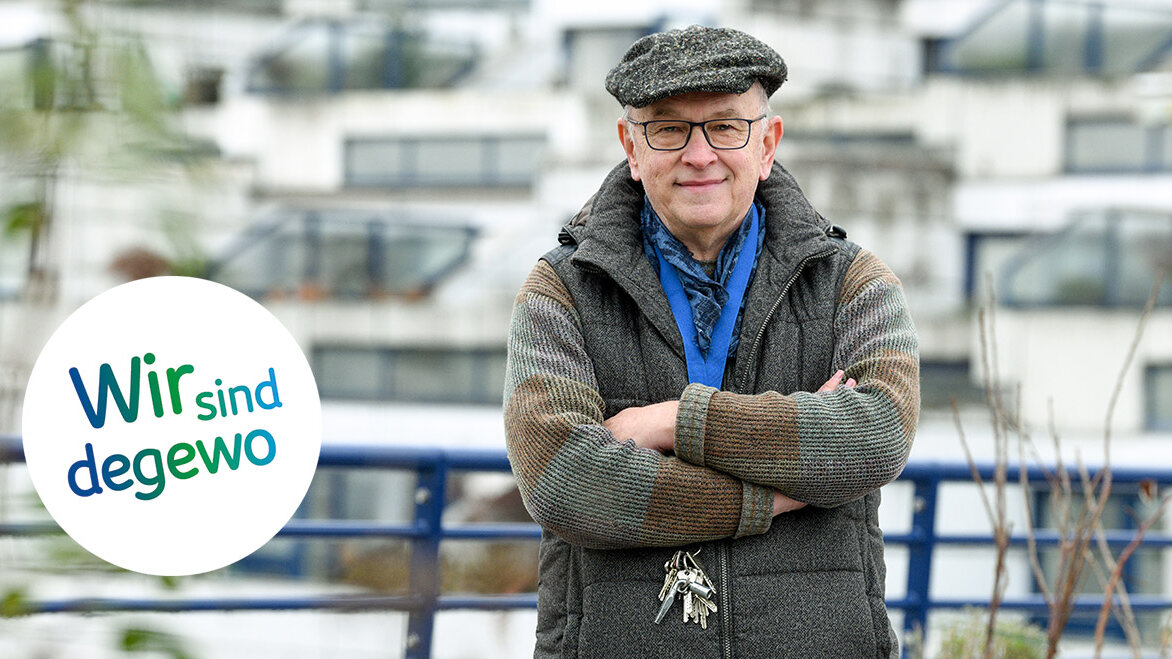 Alt-Text: Mann mit Mütze und Brille steht mit verschränkten Armen auf einem Balkon, hinter ihm ein großes Wohngebäude; links im Bild das Logo „Wir sind degewo“.