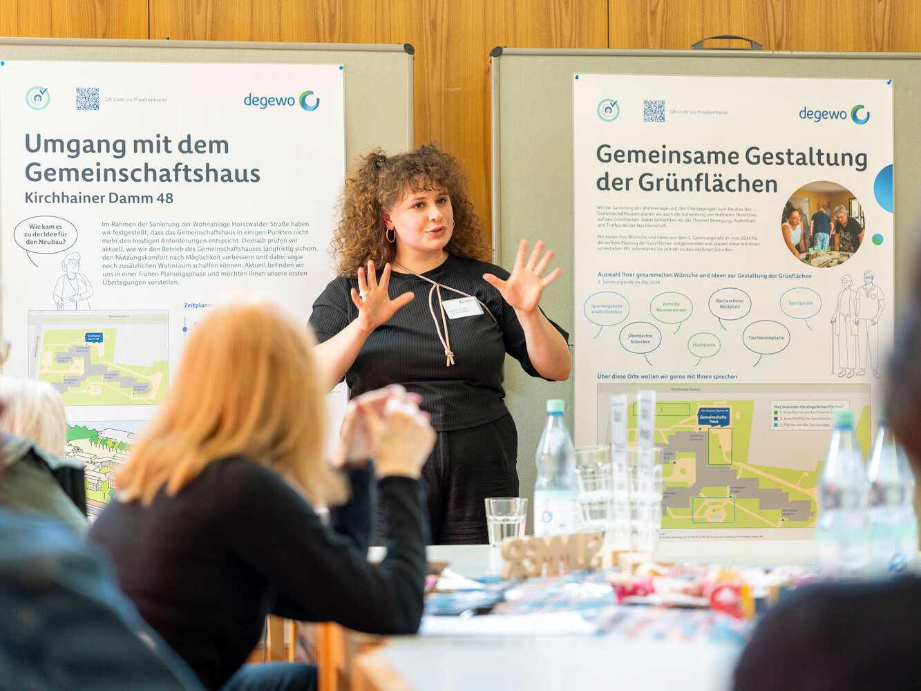 Eine Projektmitarbeiterin erläutert Inhalte zur Grünflächengestaltung vor einer Infotafel, während Bewohnende an Tischen zuhören und Fragen stellen.