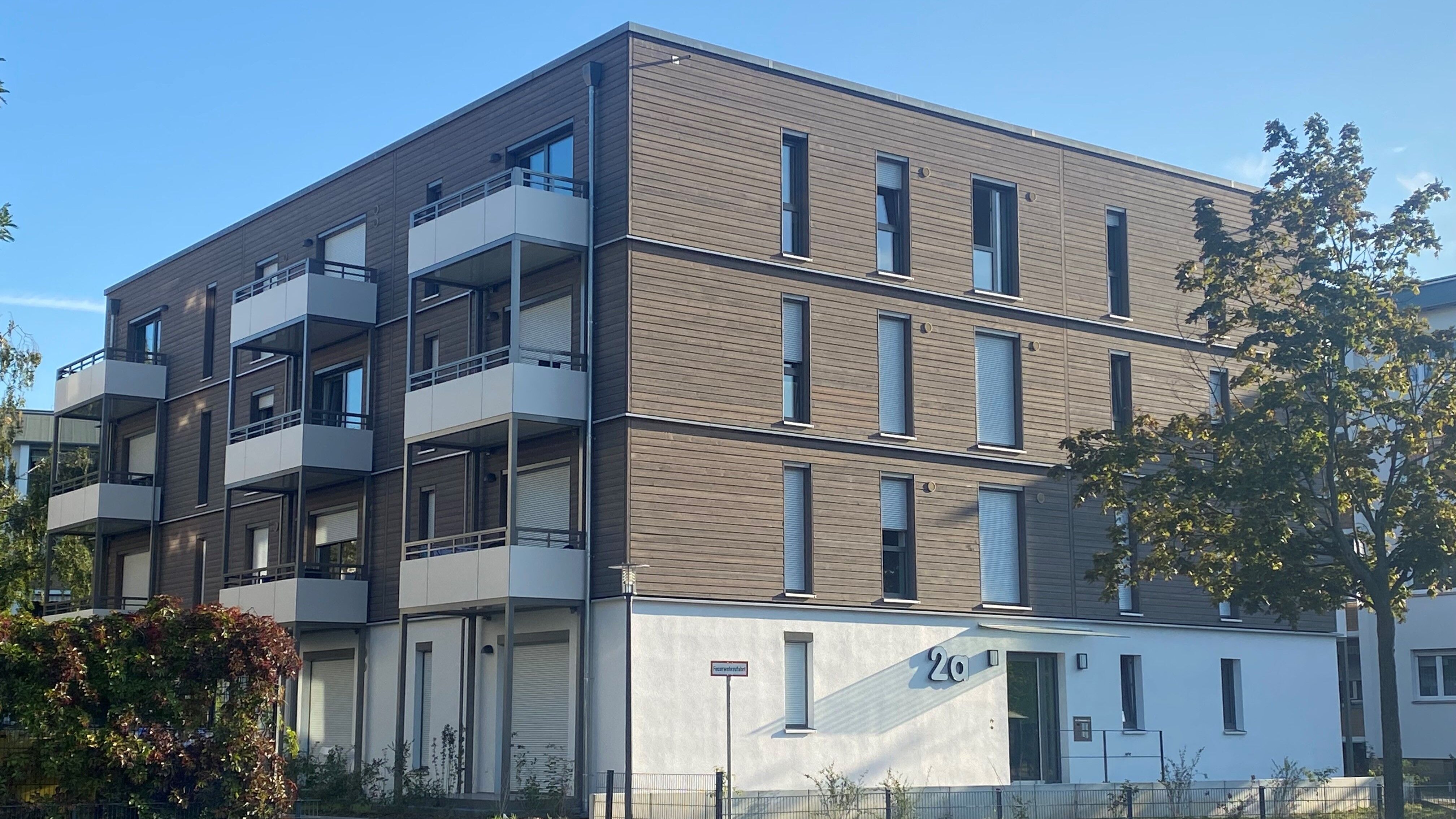 Mehrgeschossiges Wohnhaus mit moderner Holzfassade, weißen Balkonen und großen Fenstern in einem grünen Wohnquartier mit zeitgemäßer Architektur und nachhaltigem Anspruch.