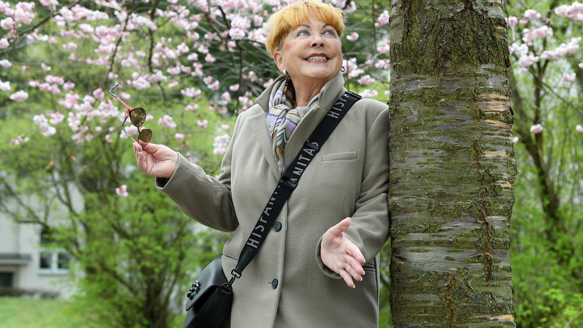 Eine ältere Frau steht lächelnd neben einem Kirschblütenbaum. Sie hält eine Sonnenbrille in der Hand schaut den Baum hinauf.