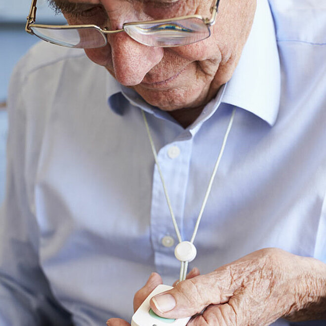 Ein älterer Herr schaut auf sein Band mit dem Notrufknopf, welches um seinen Hals hängt.
