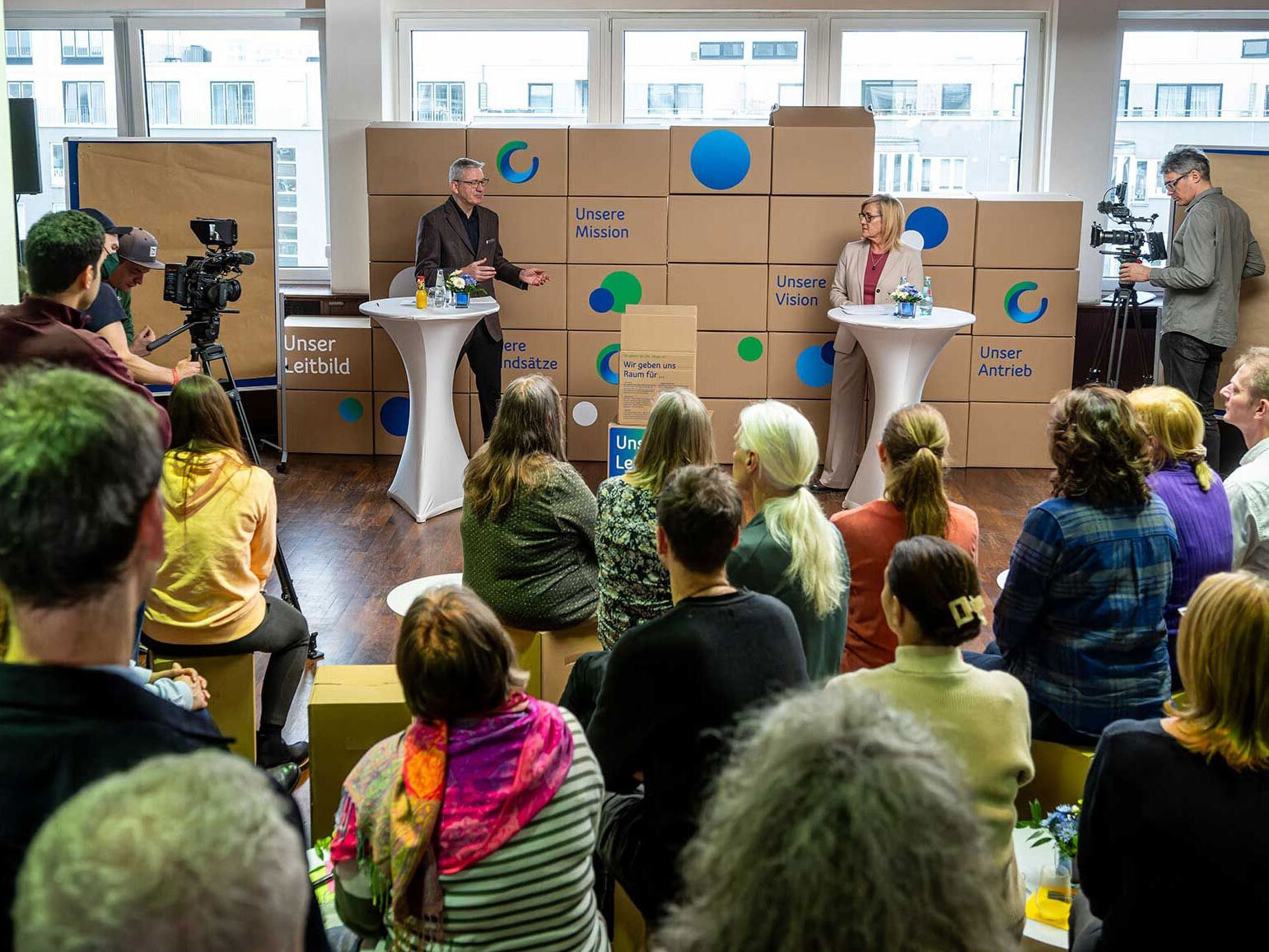 Personen sitzen in einem Veranstaltungsraum und blicken auf zwei Redner, die an Stehtischen vor einer Wand aus gestapelten Kartons stehen. Auf den Kartons sind Begriffe wie „Unsere Mission“, „Unsere Vision“, „Unser Leitbild“ und „Unser Antrieb“ zu lesen. Kamerateams filmen die Veranstaltung. Große Fenster im Hintergrund lassen Tageslicht in den Raum.
