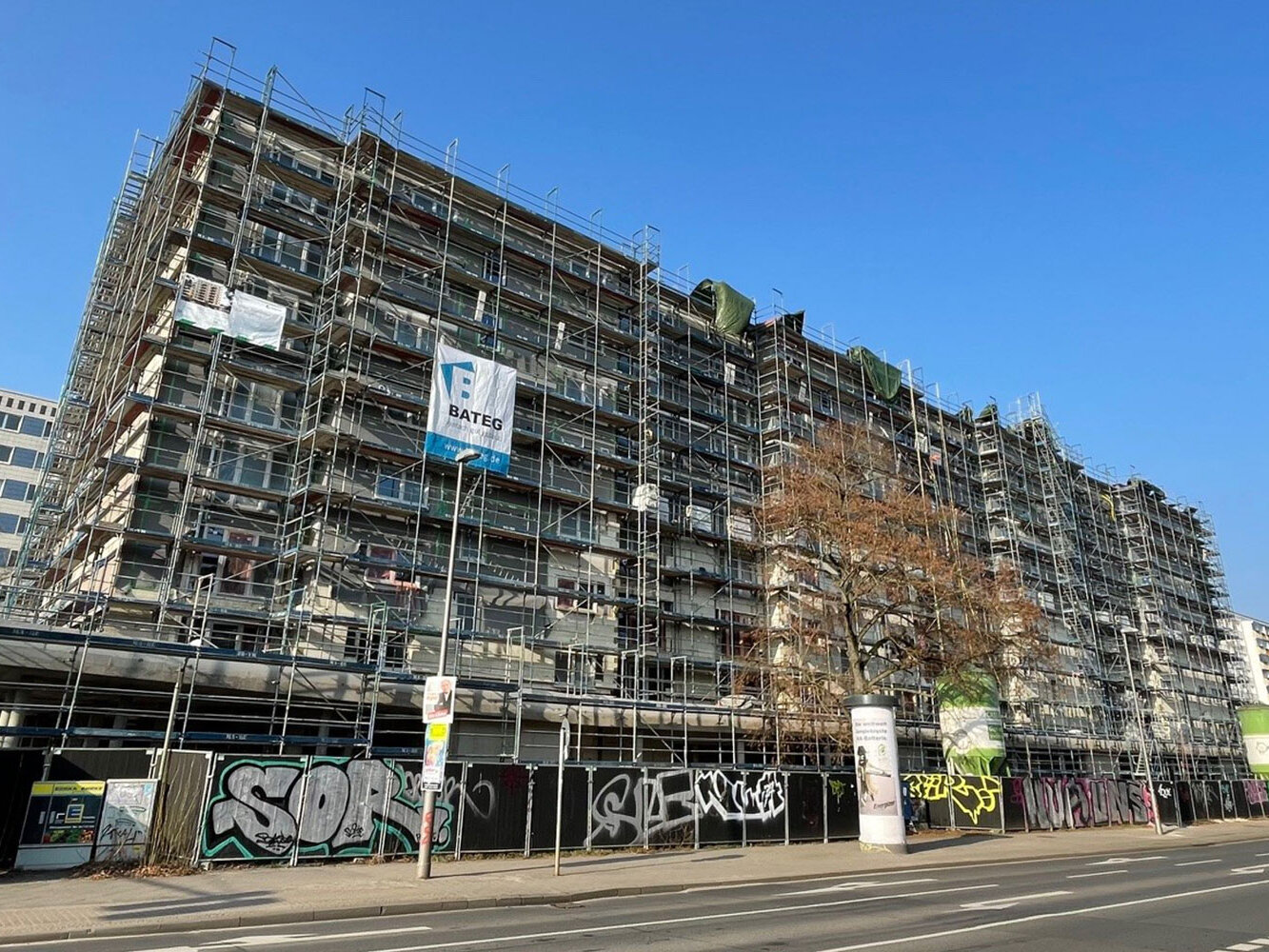Ein großes mehrgeschossiges Wohngebäude ist vollständig eingerüstet. Bauplanen und ein Banner mit der Aufschrift „BATEG“ hängen am Gerüst. Im Vordergrund verläuft eine Straße mit Gehweg und Bauzaun, der mit Graffiti besprüht ist. Der Himmel ist klar.