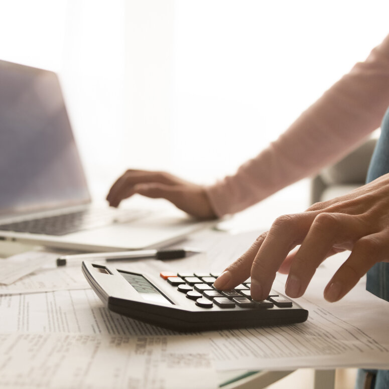 Eine Person tippt mit der linken Hand auf einen Taschenrechner, mit der anderen scrollt sie auf einem Laptop. 