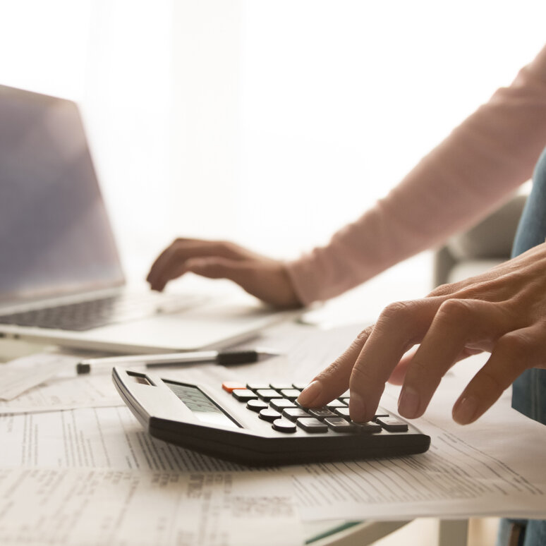 Eine Person tippt mit der linken Hand auf einen Taschenrechner, mit der anderen scrollt sie auf einem Laptop. 