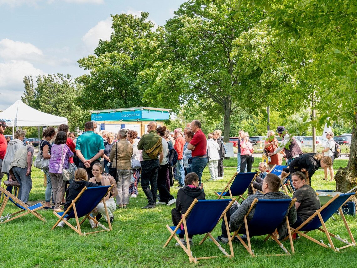 Besucherinnen und Besucher informieren sich im Park an der Dialogstation über das Neubauvorhaben; Liegestühle laden zum Verweilen ein.