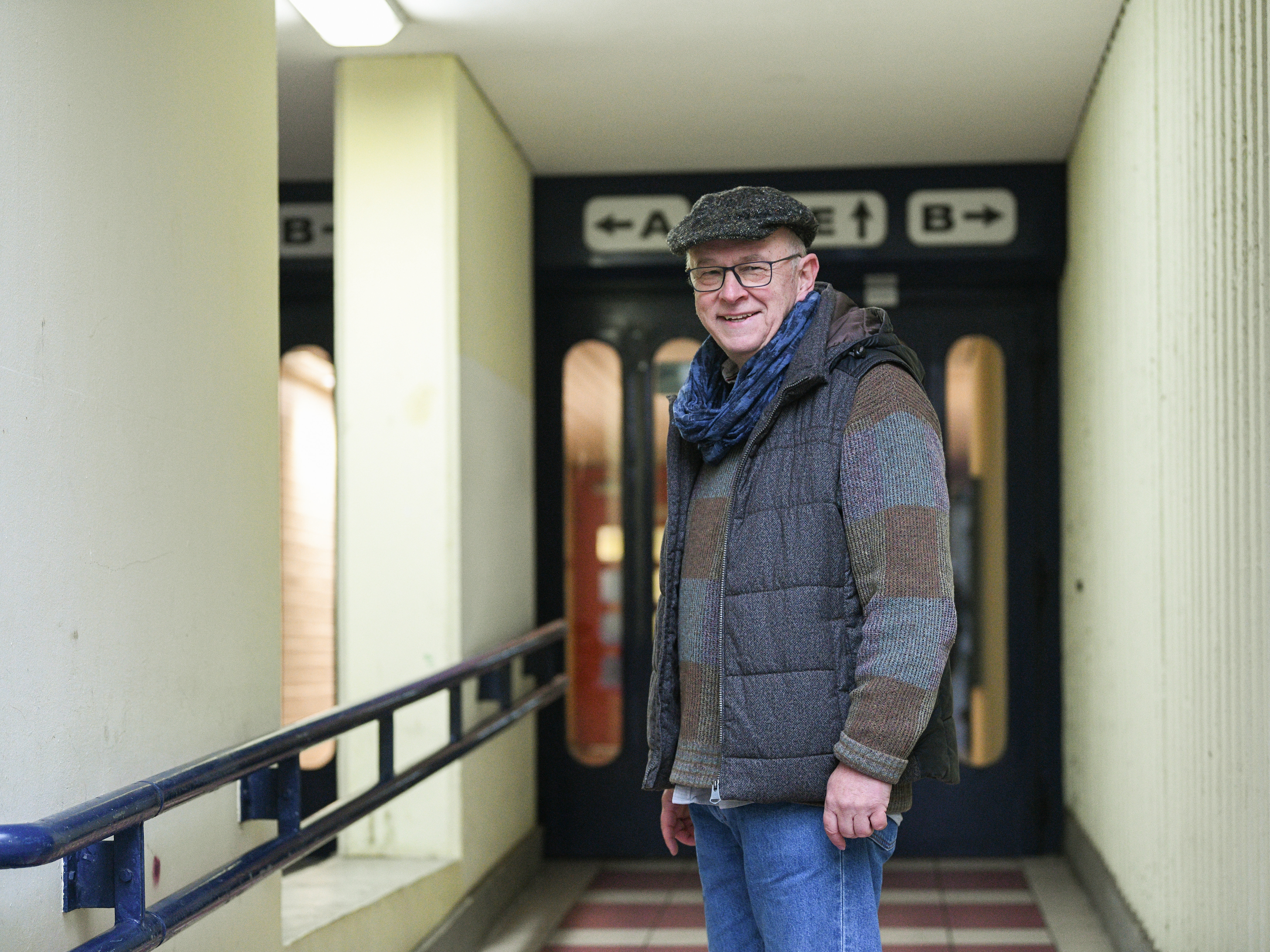 Mann mit Mütze, Brille und Schal steht in einem Hauseingang mit Rampe und schaut lächelnd in die Kamera.