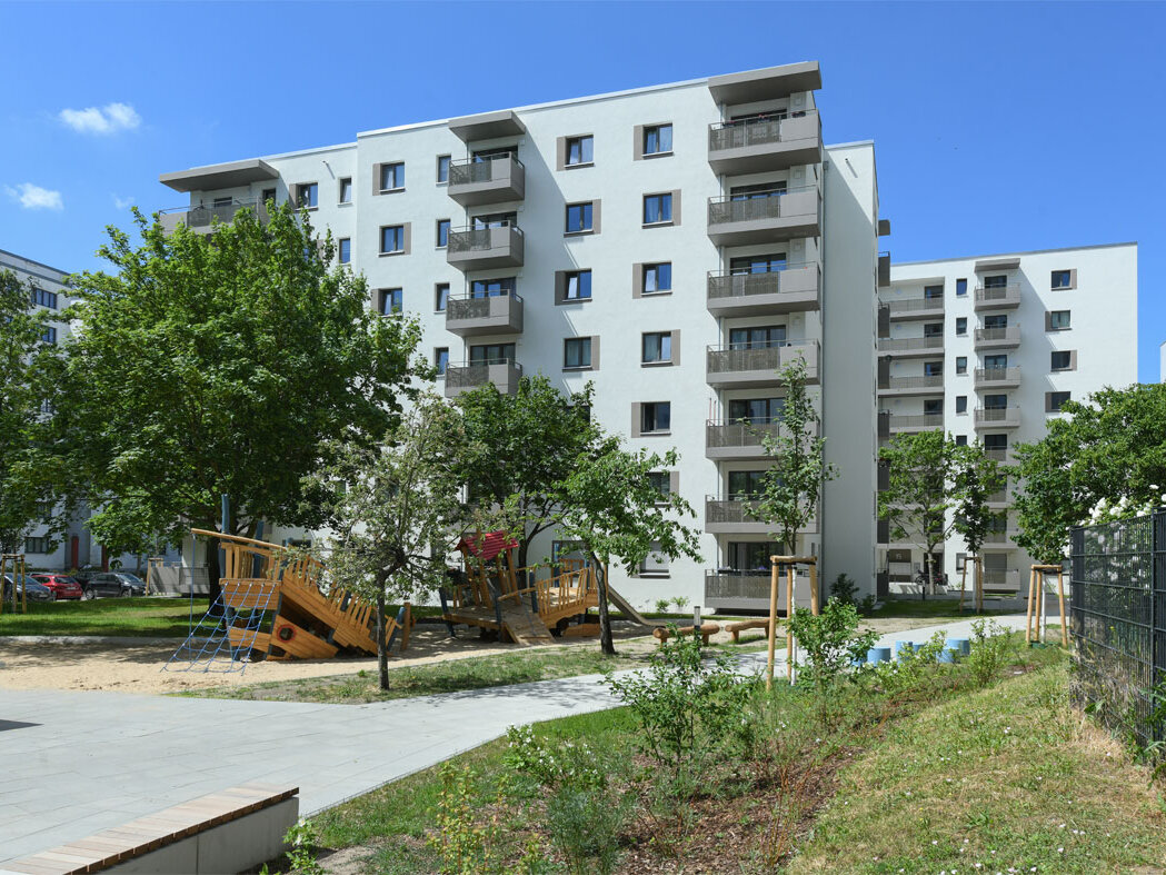 Außenansicht degewo-Neubau Theodor-Loos-Weg 15 und 15 A mit Weg, Sitzbank, Beeten, Rasenhang und Zaun im Vordergrund.