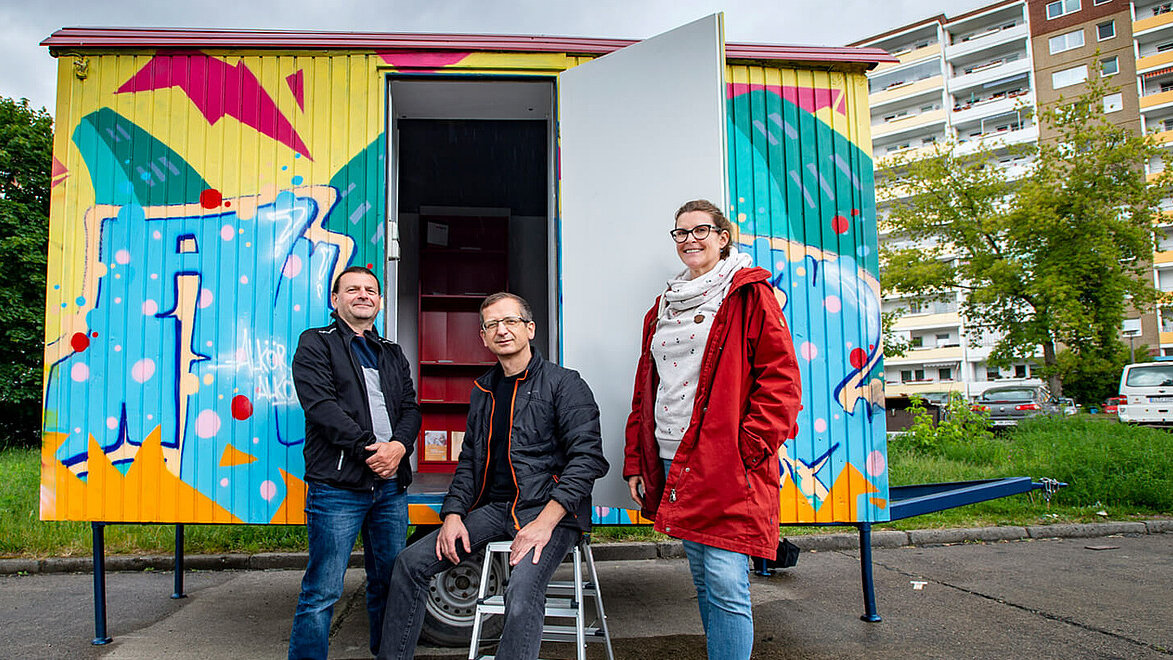 Drei Personen stehen vor einem bemalten Container.