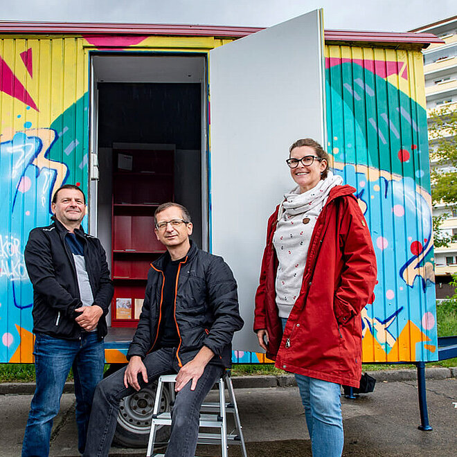 Drei Personen stehen vor einem bemalten Container.