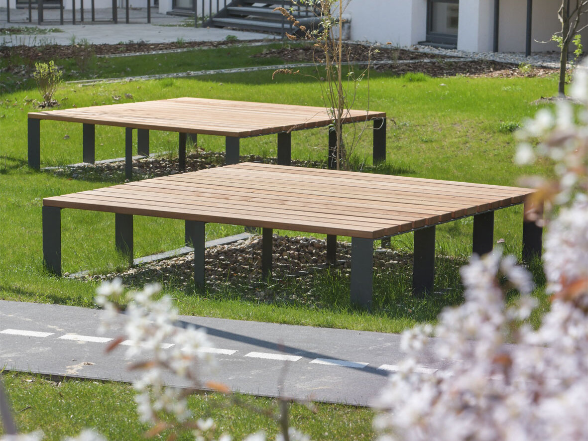 Außenansicht degewo-Neubau Lützowstraße 7: Rasenfläche mit Sitzgelegenheiten und neu gepflanzten Büschen und Bäumen, im Vordergrund blühende Zweige.