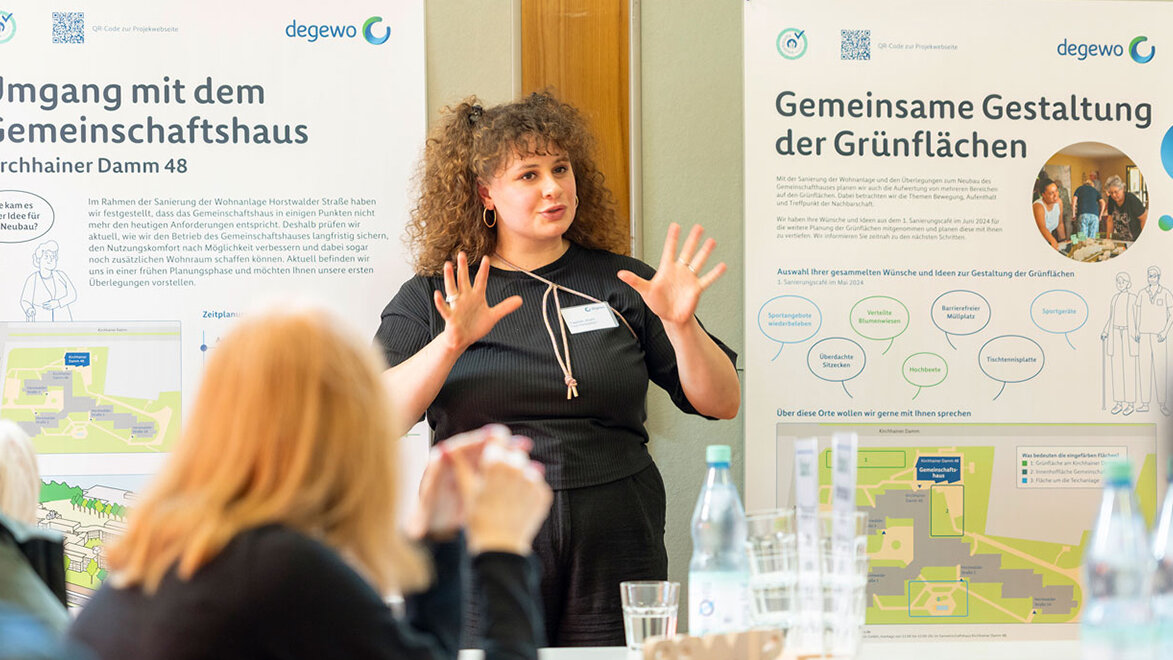 Eine Referentin präsentiert vor Plakaten zur gemeinsamen Gestaltung der Grünflächen. Mehrere Personen hören aufmerksam zu.