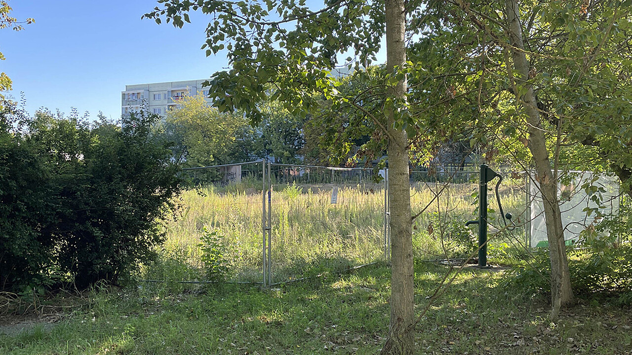 Grundstück in der Wuhlestraße, dass für den Neubau vorgesehen ist. Es ist ein Bauzaun auf einer Wiese zu sehen, dahinter ein Gebäude.