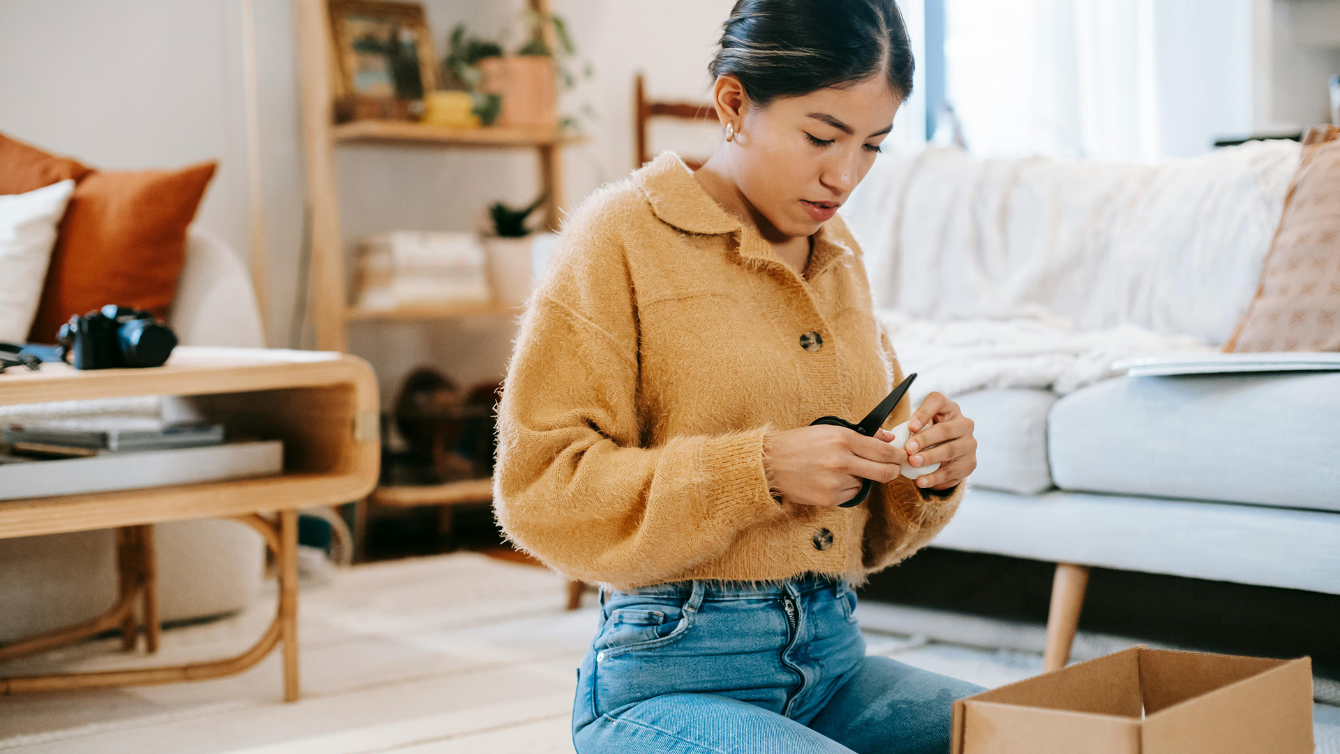 Person sitzt in einer Wohnung auf dem Boden und öffnet mit einer Schere einen Karton. Im Hintergrund sind Sofa, Regal und Wohnaccessoires zu sehen.