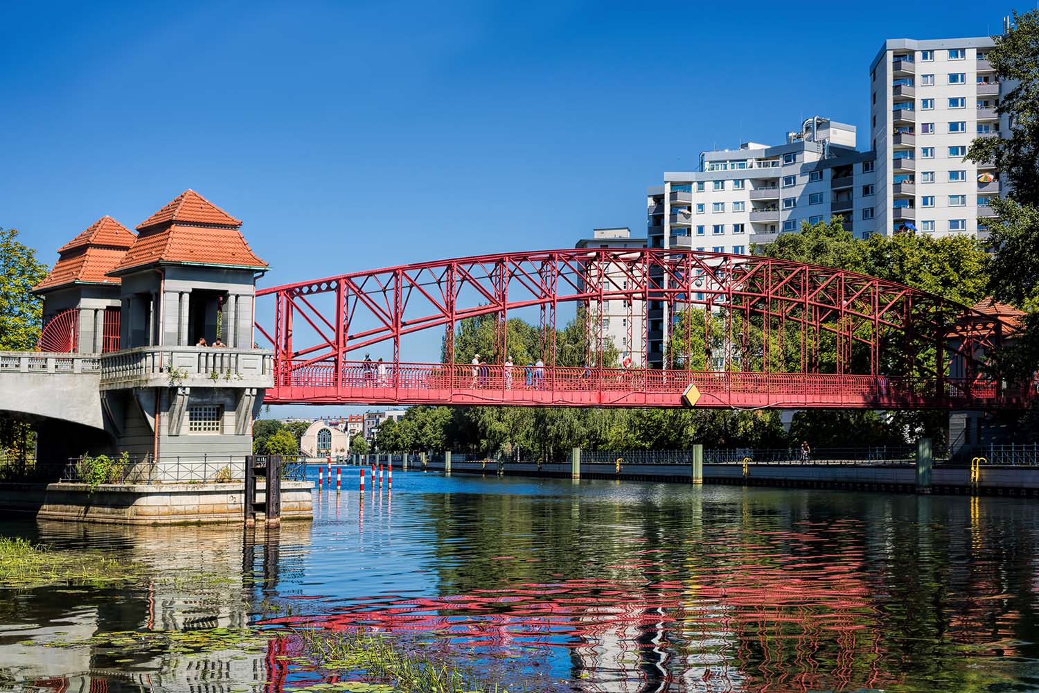 Rote Stahlbrücke über den Tegeler Hafen mit Wasserkanal und Wohngebäuden im Hintergrund.