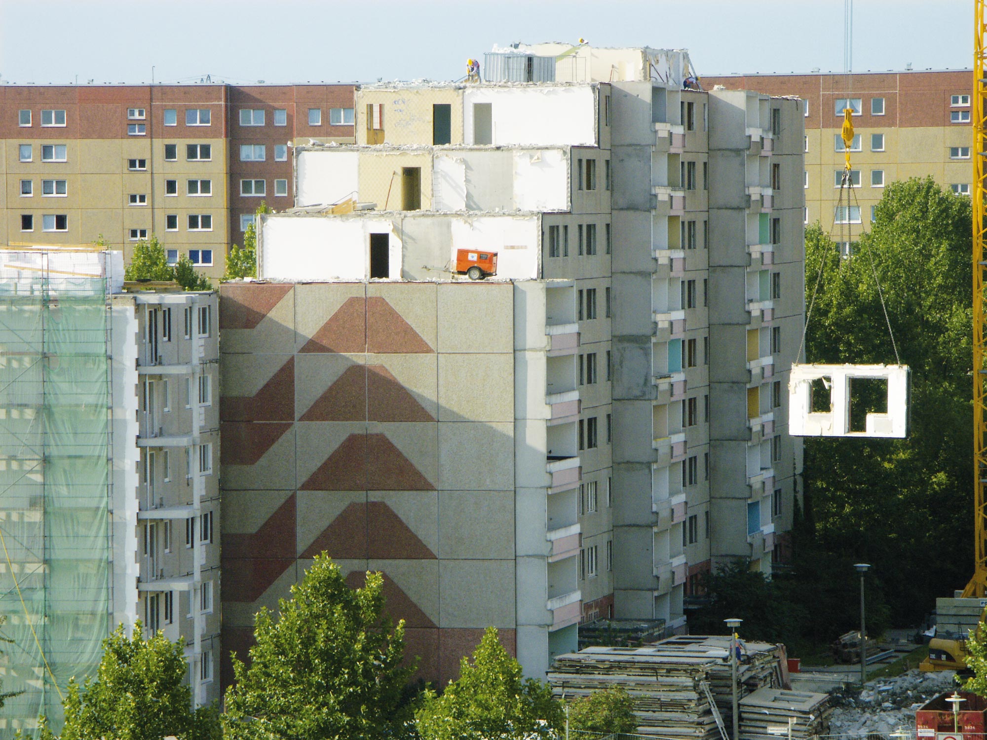 Mehrgeschossiges Wohngebäude im Rückbau, bei dem mehrere obere Etagen bereits entfernt wurden; ein Kran hebt ein Bauelement ab, im Hintergrund weitere Wohnhäuser.