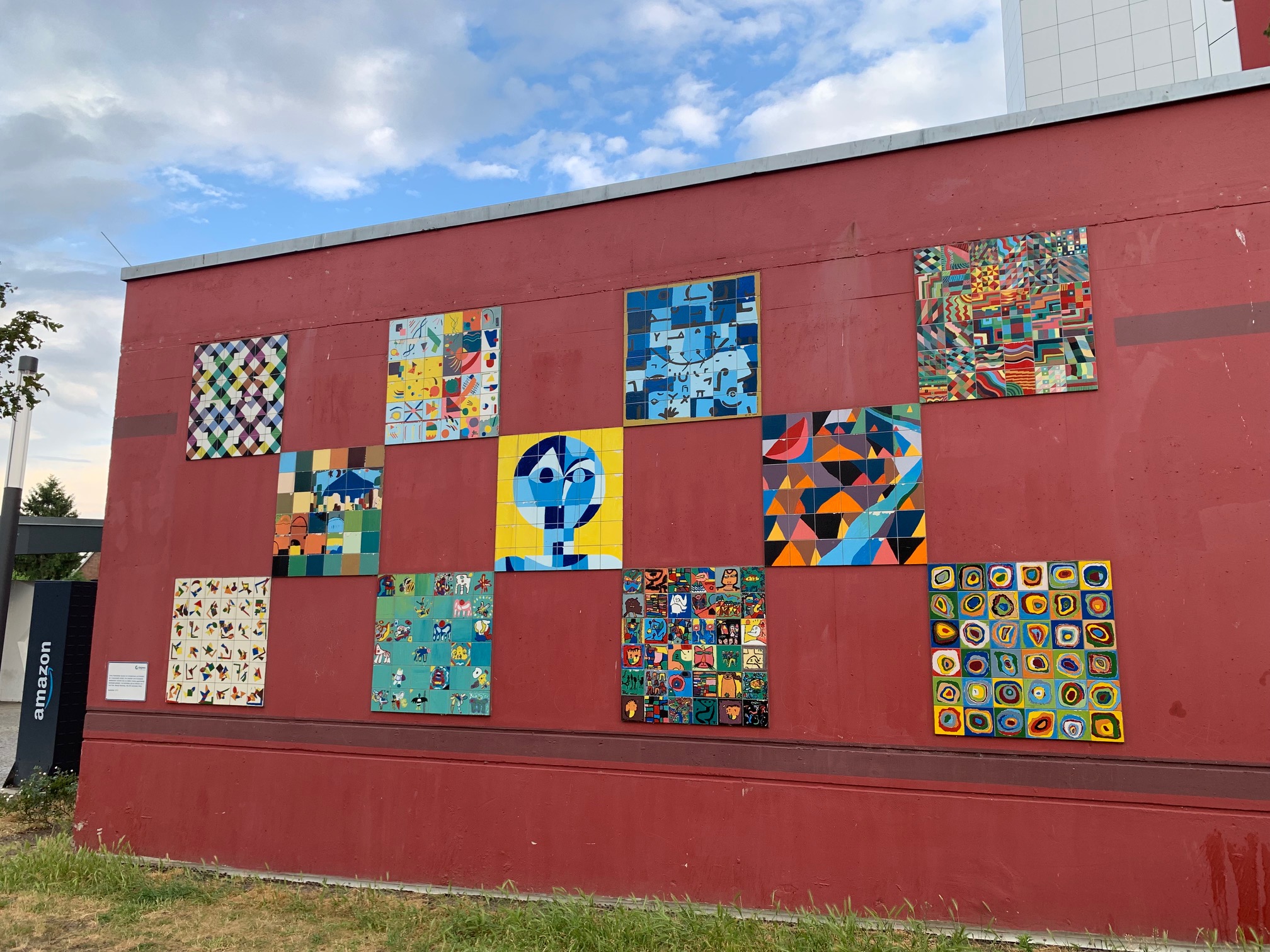 Elf verschiedene, quadratische Wandbilder in bunten Farben auf einer dunkelroten Wand.