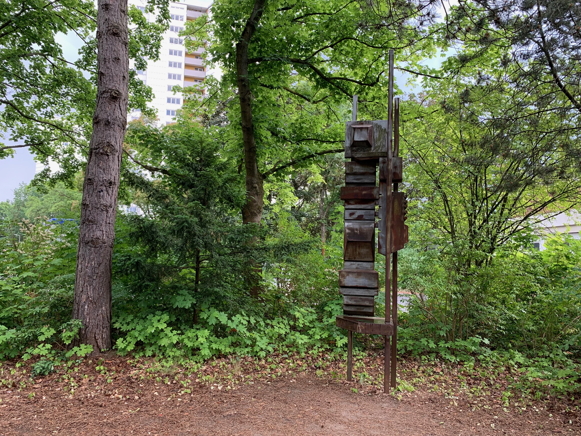 Eine abstrakte Skulptur aus teilweise rostigem Stahl die vor grünen Bäumen und Sträuchern steht.