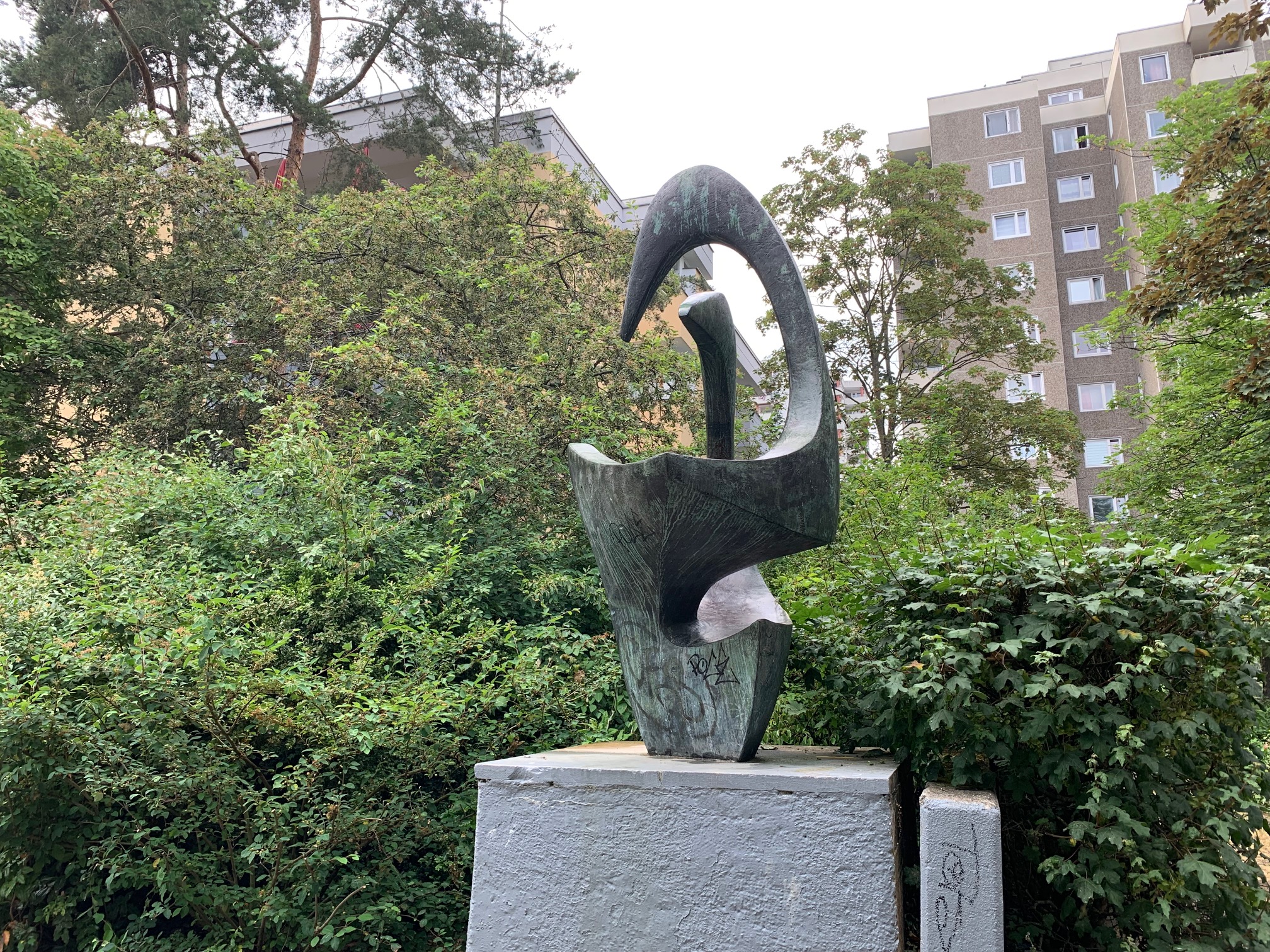 Eine abstrakte Skulptur aus oxidierter Bronze auf einem Betonsockel vor grünen Sträuchern. Im Hintergrund hohe Wohnhäuser.