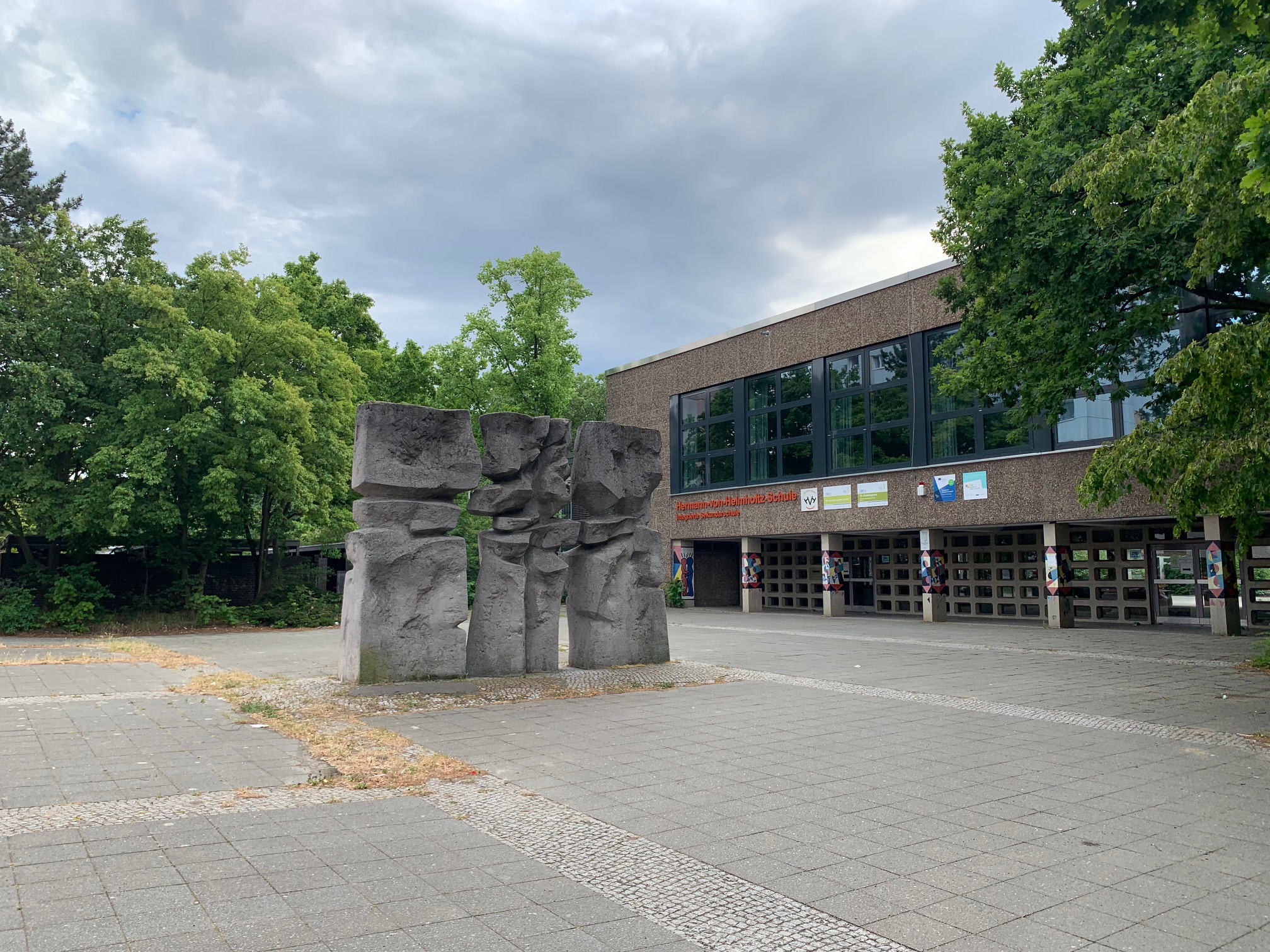 Eine Steinmauer die durch unregelmäßig geformte Löcher im Material den Eindruck erweckt, zerbrochen zu sein. Das Objekt befindet sich im Eingangsbereich eines Schulgebäudes.