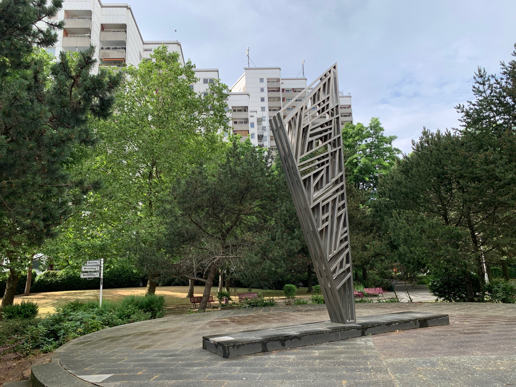 Eine abstrakte Stahlskulptur mit vielen unterschiedlich angeordneten Streben auf einem runden, flachen Podest, das begehbar ist. Im Hintergrund Bäume und hohe Wohnhäuser.