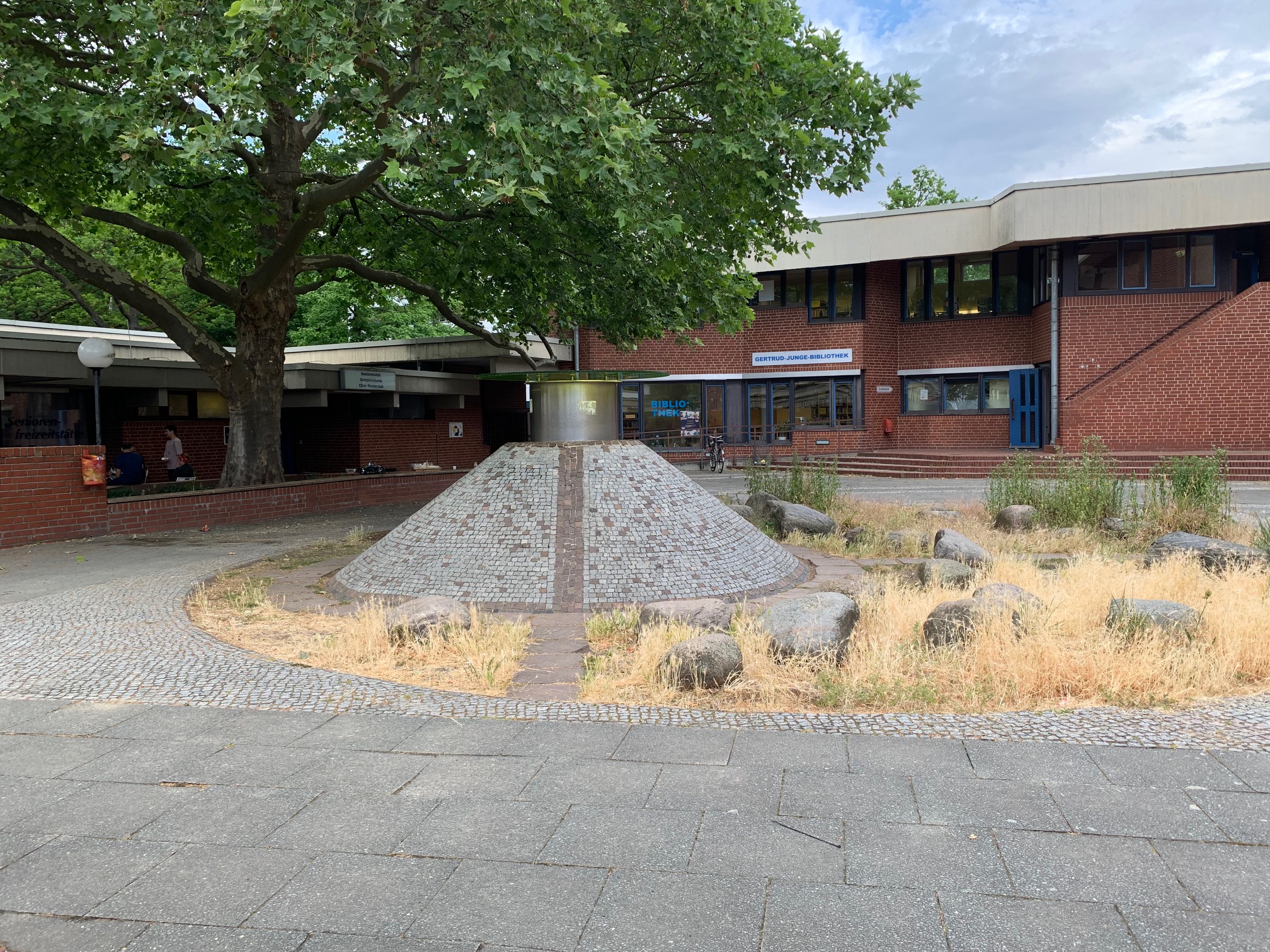 Eine kegelförmige Skulptur, die mit hellen Ziegeln verkleidet ist. Auf der Spitze ein zylinderförmiger Aufbau aus einem anderen Material. Im Hintergrund ein Gebäude und ein Baum.