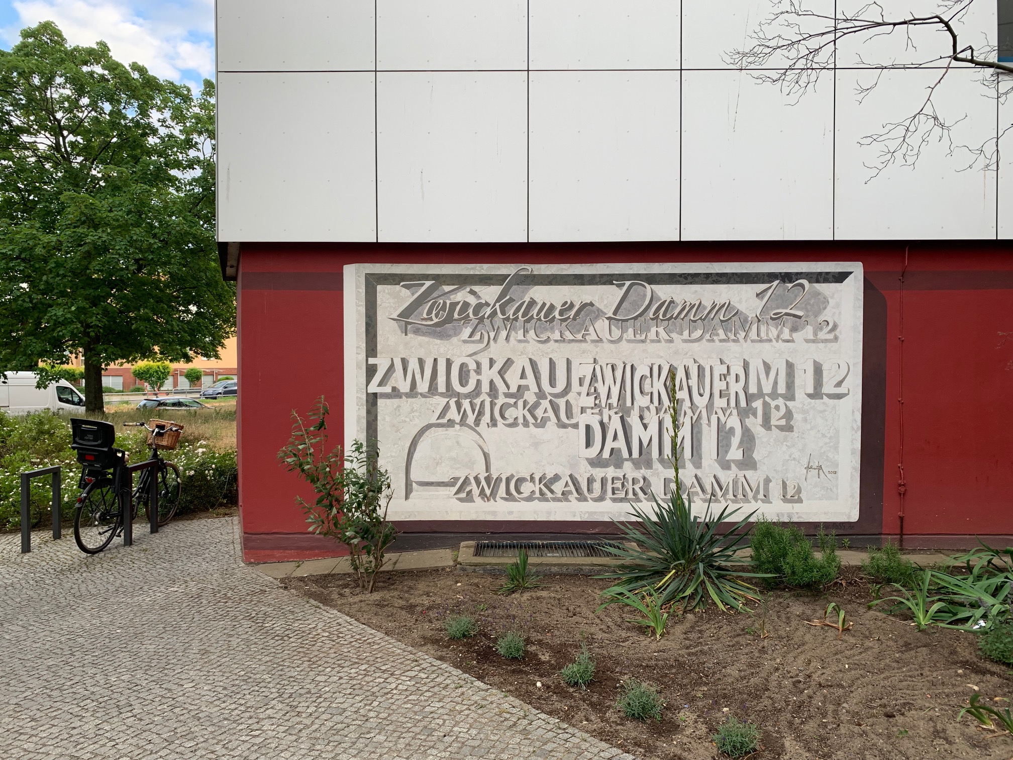 Ein rechteckiges, grau-weißes Wandgemälde auf dunkelrotem Untergrund, in dem mehrfach in verschiedenen Schriftarten „Zwickauer Damm 12“ geschrieben wurde.