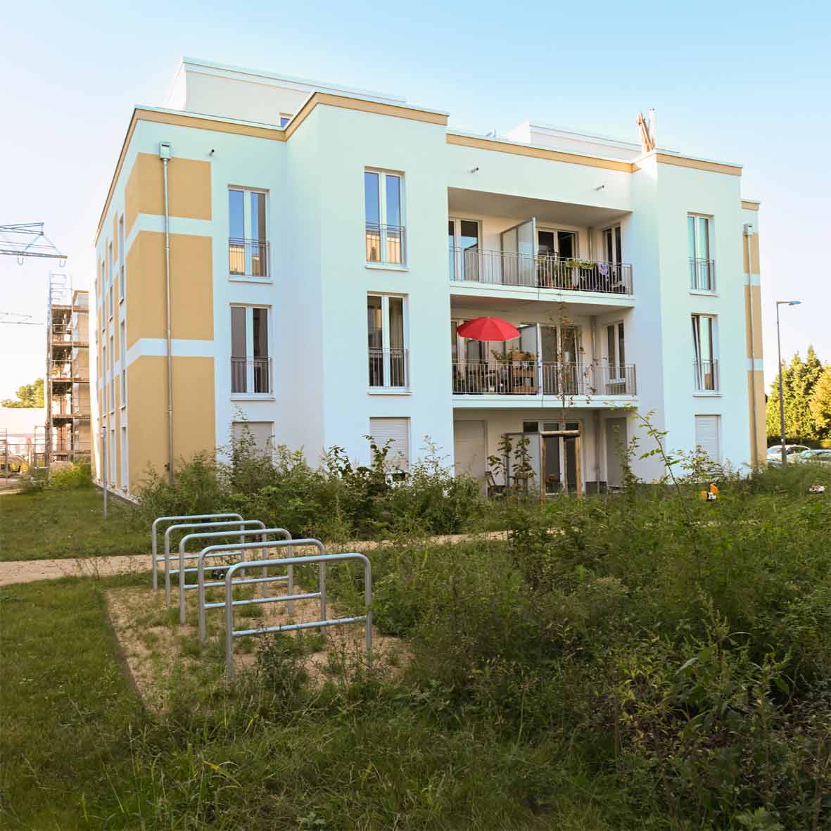 Schrägansicht eines degewo-Neubaus am Eisenhutweg mit drei Vollgeschossen und Staffelgeschoss. Die Fassade ist hell gestaltet und mit gelben Akzentflächen gegliedert. Balkone mit Metallgeländern öffnen sich zur begrünten Außenanlage; im Vordergrund sind Fahrradständer und Rasenflächen zu sehen