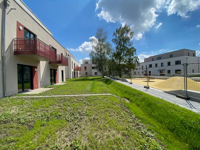Außenansicht degewo-Neubau Fürstenwalder Allee 324/326: links Gebäude mit Grünfläche davor, rechts Spielplatz mit Bauzaun, im Hintergrund Bäume und weitere Gebäude.