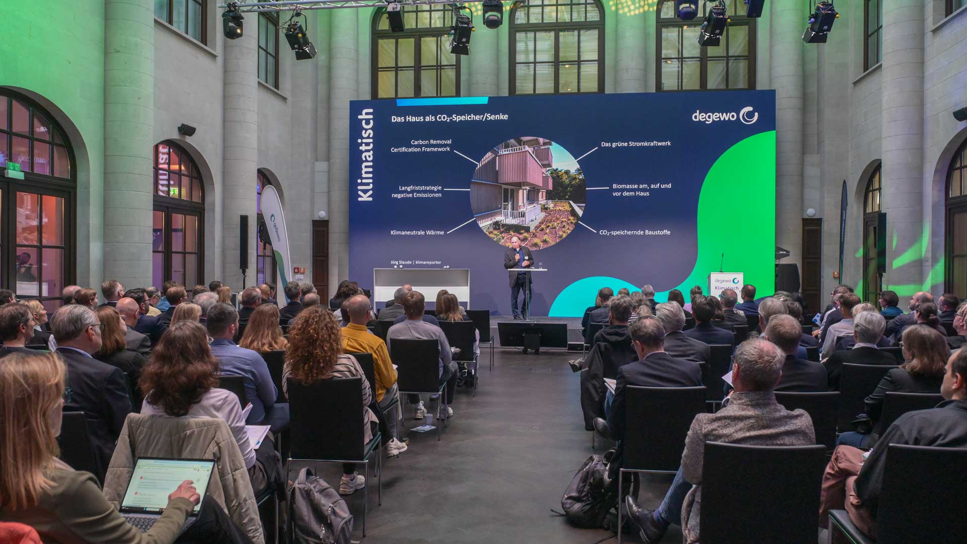 Copilot said: Alt-Text (Web, kurz): Publikum bei einer Fachveranstaltung in einer großen Halle. Auf der Bühne präsentiert eine Person vor einer großen Projektion zum Thema „Das Haus als CO₂-Speicher/Senke“ im Rahmen der degewo-Veranstaltung „Klimatisch“.