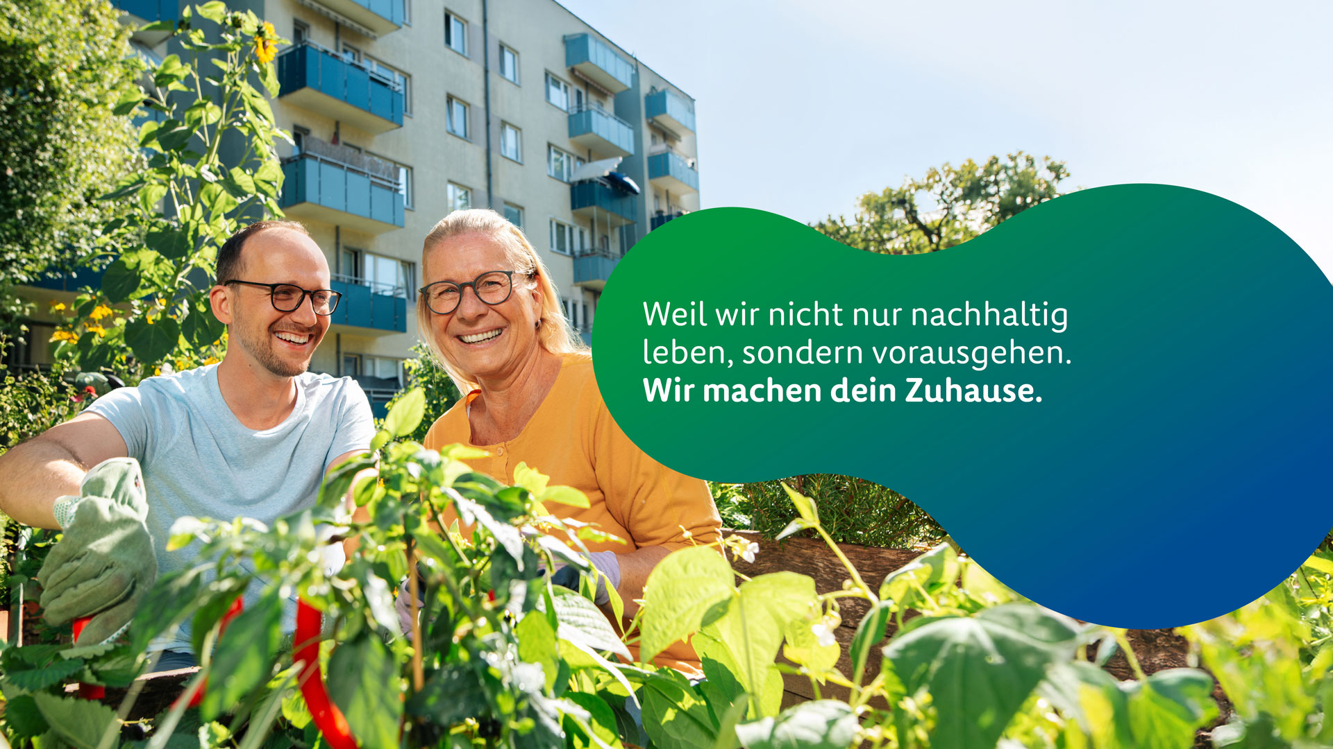 Zwei Personen sitzen in einem begrünten Innenhof vor einem mehrgeschossigen Wohngebäude mit Balkonen und bepflanzen gemeinsam ein Hochbeet. Beide tragen Handschuhe und lächeln in die Kamera. Im Vordergrund sind grüne Pflanzen und Blätter zu sehen. Rechts im Bild befindet sich eine große grün-blaue Grafikfläche mit dem Schriftzug: „Weil wir nicht nur nachhaltig leben, sondern vorausgehen. Wir machen dein Zuhause.“