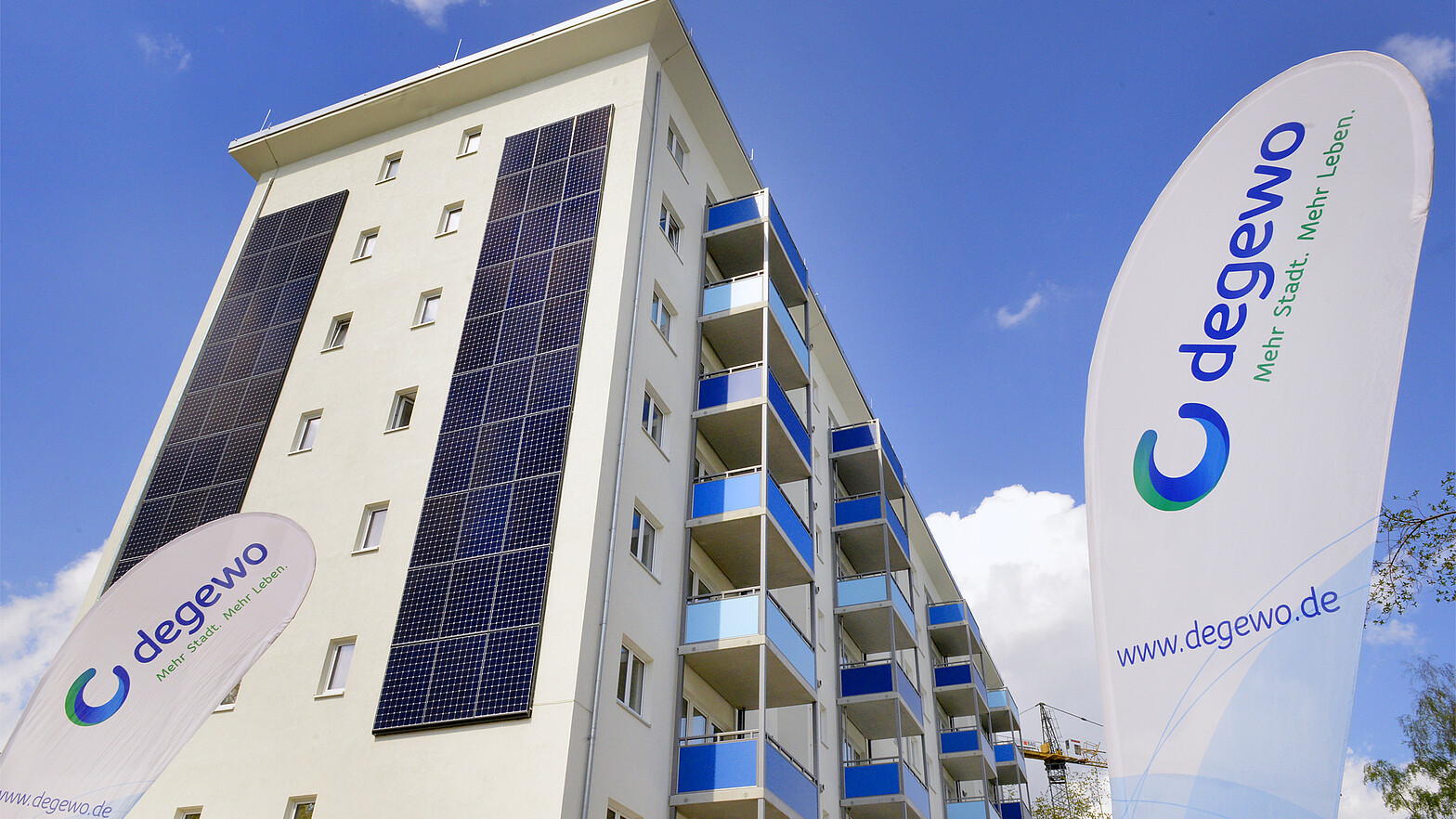 Von unten Blick nach oben auf ein großes Mietshaus mit Solarpaneelen an den Außenwänden und Flaggen von degewo davor